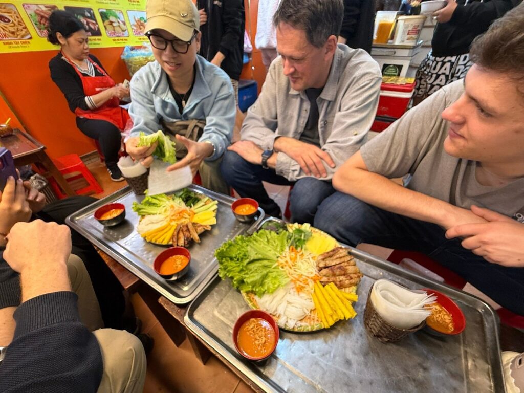 Diner assembling a rice-paper roll while others watch beside platters of herbs and grilled meat.