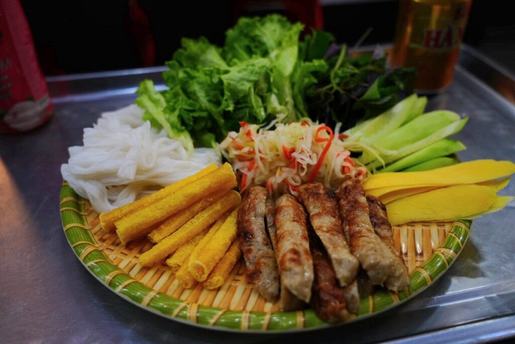 Platter of grilled meat rolls, rice noodles, herbs, cucumber and mango on a woven tray.