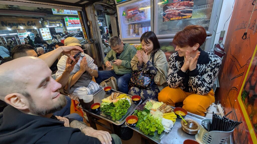 Diners gathered around platters of grilled meat, rice noodles, herbs and dipping sauces.