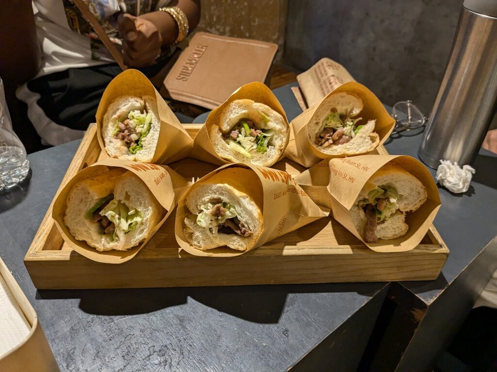Six halves of banh mi with meat and vegetables arranged on a wooden tray.