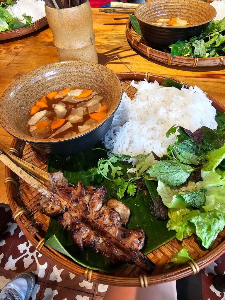 Grilled pork, rice noodles, fresh herbs and dipping sauce arranged on a woven tray.