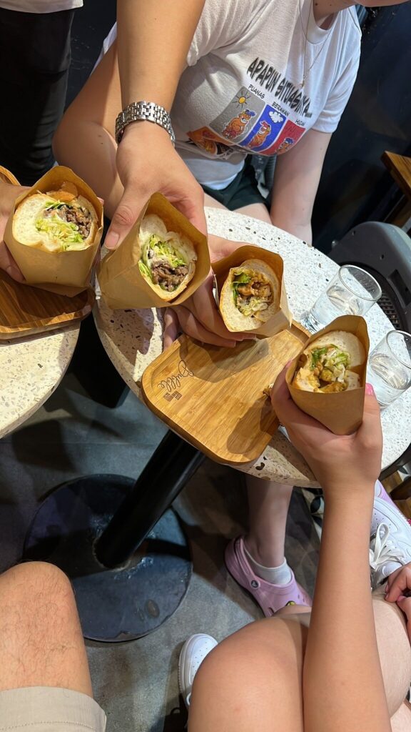 Four people holding banh mi filled with meat and vegetables above a cafe table.