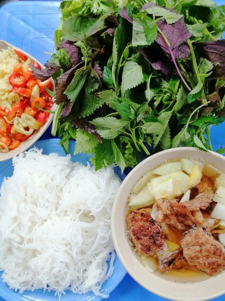 Bun cha with grilled pork, rice noodles, fresh herbs, garlic and chillies on a blue table.