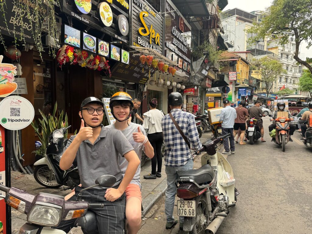 A rider and helmeted passenger giving thumbs-up on a motorbike in Hanoi's busy Old Quarter.