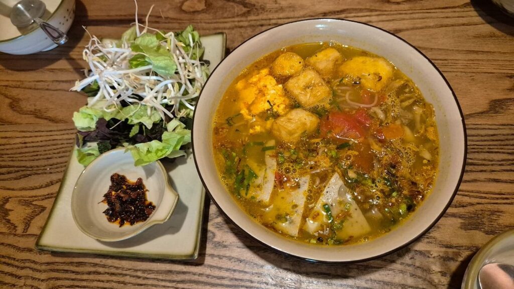 A bowl of noodle soup with tofu, tomatoes and herbs beside fresh greens.