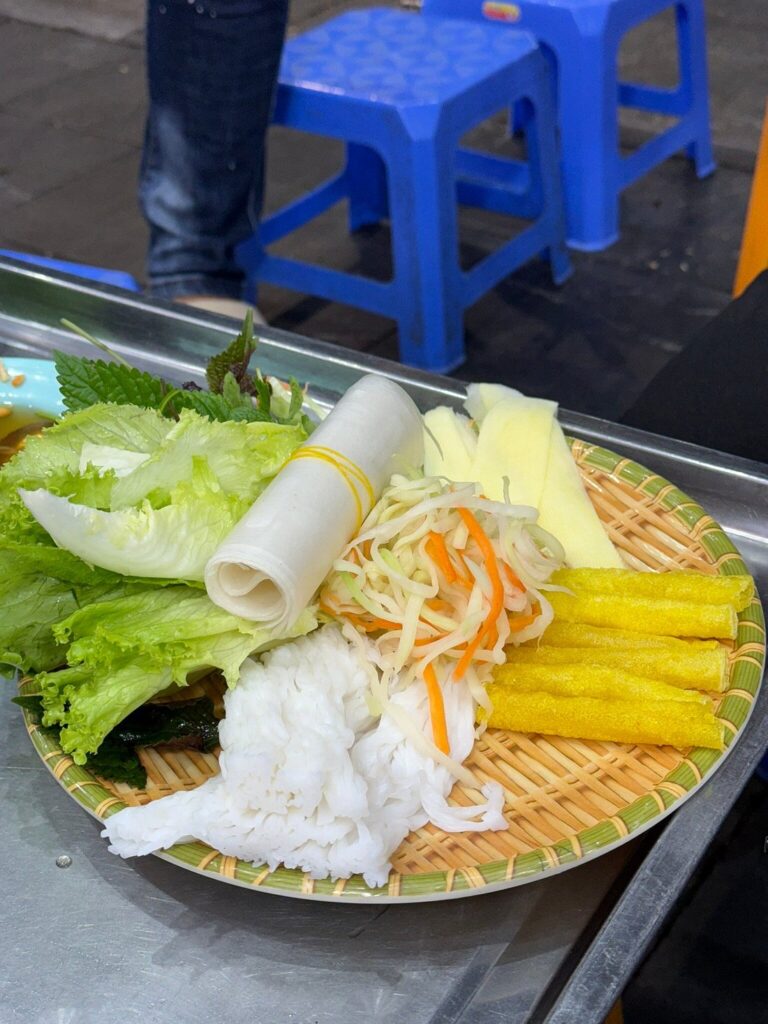 Rice paper, vermicelli, fresh herbs and sliced vegetables on a woven tray.