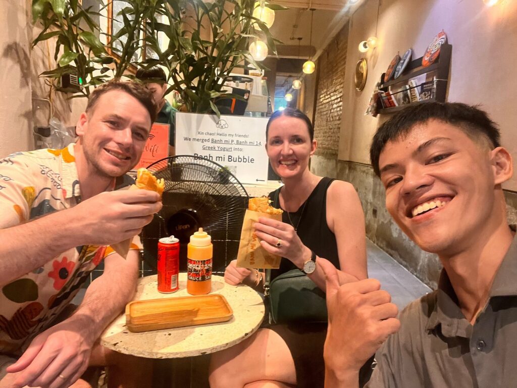 Three people smiling with banh mi sandwiches in a Hanoi eatery.