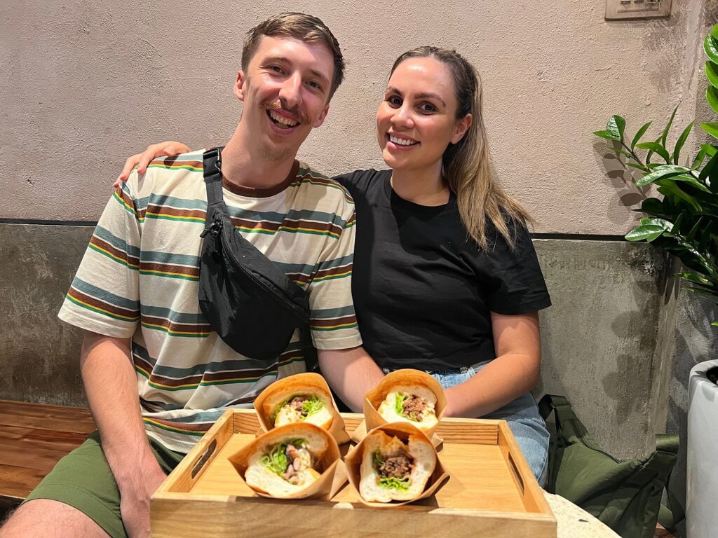 Two diners smiling behind a tray of banh mi sandwiches in Hanoi.