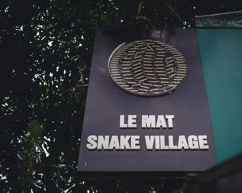 Le Mat Snake Village sign displayed beneath leafy trees.